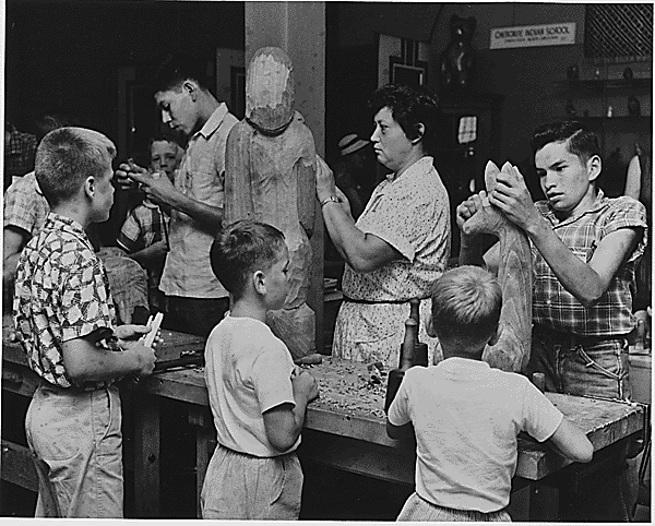 Visitors to the Craftsmen's Fair at the Cherokee Indian Reservation, Cherokee North Carolina, watch Amanda Crowe, Well-Known Cherokee Wood Scultptor and Her Students Demonstrate Their Art.