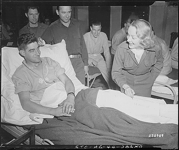 Marlene Dietrich, motion picture actress, autographs the cast on the leg of Tec 4 Earl E. McFarland of Cavider, Texas, at a United States hospital in Belgium, where she has been entertaining the GIs., ca. 11/24/1944