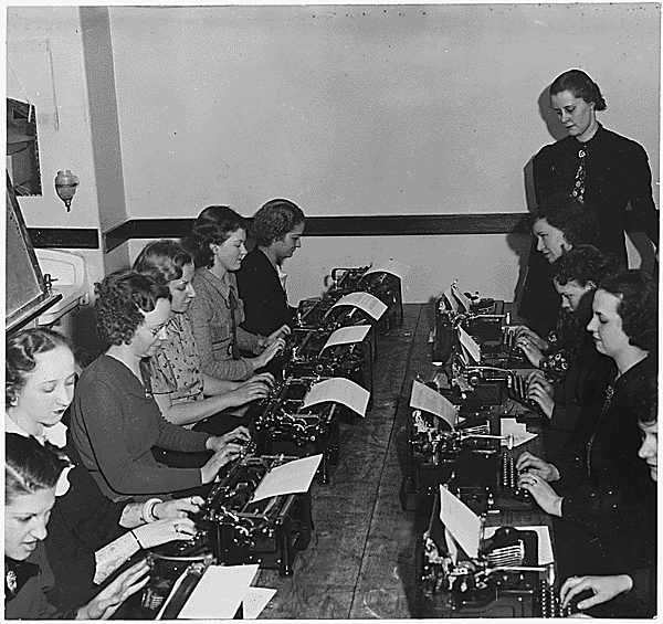 Women sitting at typewriters around a table in black and white photo